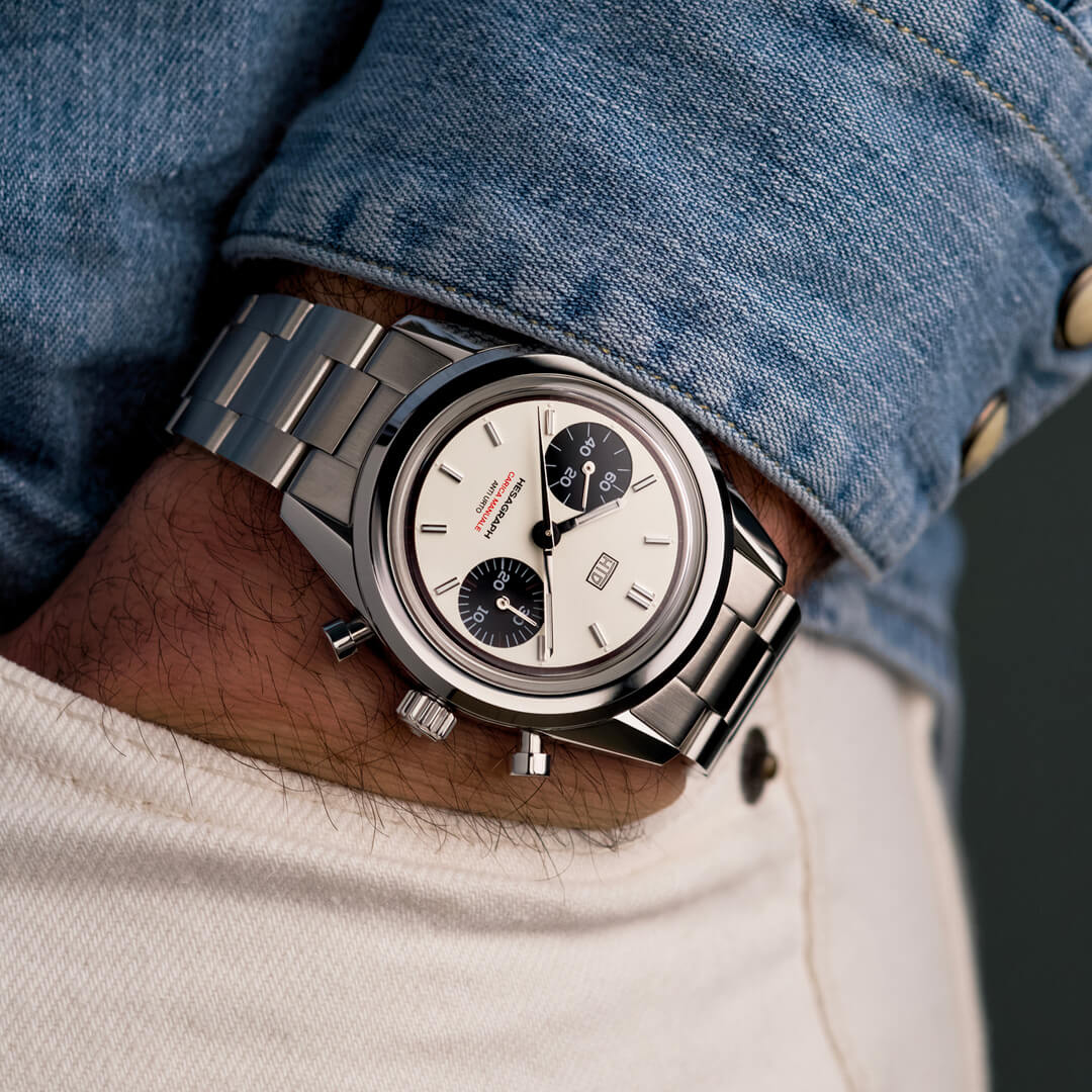 Orologio cronografo in acciaio con quadrante bianco e contatori neri, indossato al polso con camicia di jeans e pantaloni chiari.