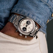 Orologio cronografo in acciaio con quadrante bianco e contatori neri, indossato al polso con camicia di jeans e pantaloni chiari.