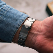 Dettaglio della chiusura in acciaio con logo HTD di un orologio da polso, indossato con camicia di jeans.