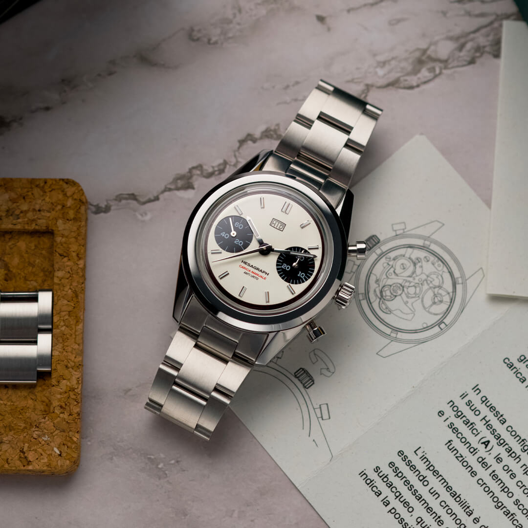 Orologio cronografo HTD Hesagraph in acciaio con quadrante bianco e contatori neri, appoggiato su un tavolo accanto a manuale illustrato e maglie di ricambio.