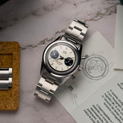 Orologio cronografo HTD Hesagraph in acciaio con quadrante bianco e contatori neri, appoggiato su un tavolo accanto a manuale illustrato e maglie di ricambio.