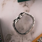 Vista laterale di un orologio cronografo in acciaio con bracciale metallico e corona incisa, appoggiato su superficie marmorea.