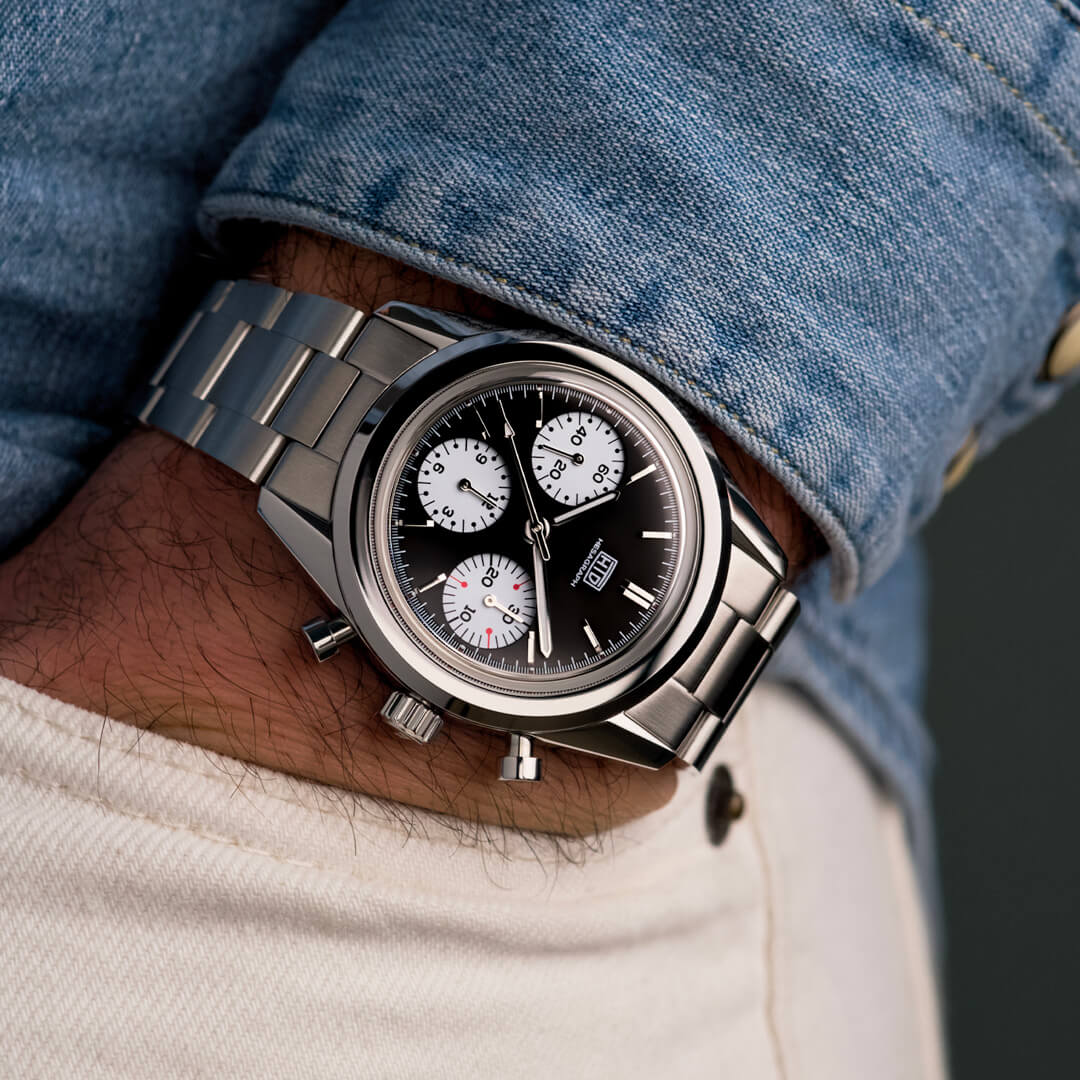 Primo piano di un orologio cronografo da polso con quadrante nero e contatori bianchi, indossato con camicia di jeans e pantaloni chiari.