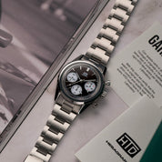 Orologio cronografo in acciaio con quadrante nero e tre contatori bianchi, appoggiato su un tavolo accanto a un libretto e alla card di garanzia HTD.