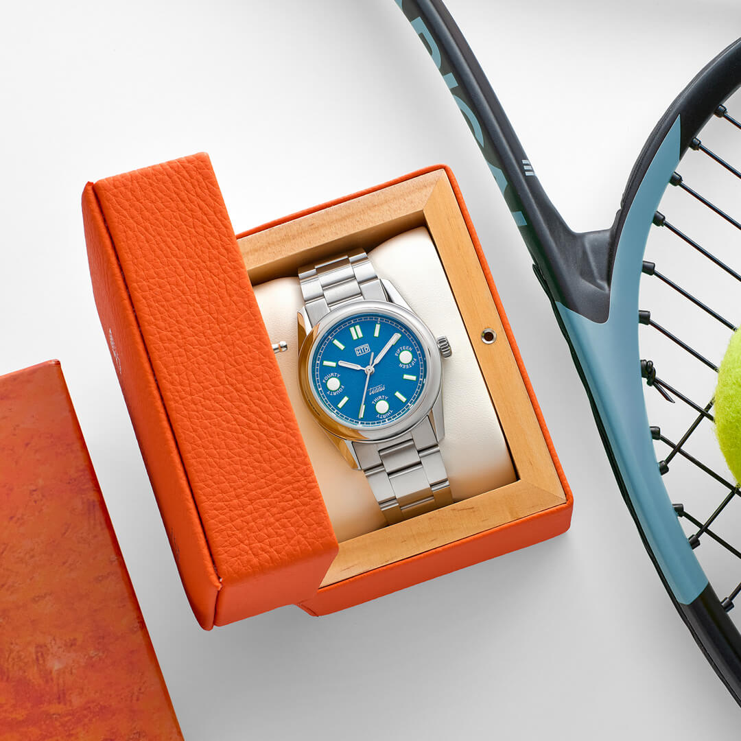Orologio HTD Tennis Cemento in esposizione nella scatola arancione, accanto a racchetta e pallina da tennis.