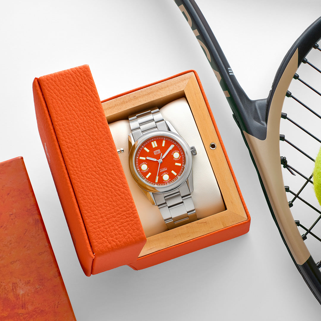 Orologio HTD Tennis Terra con quadrante arancione in esposizione all’interno della scatola arancione, accanto a una racchetta da tennis.